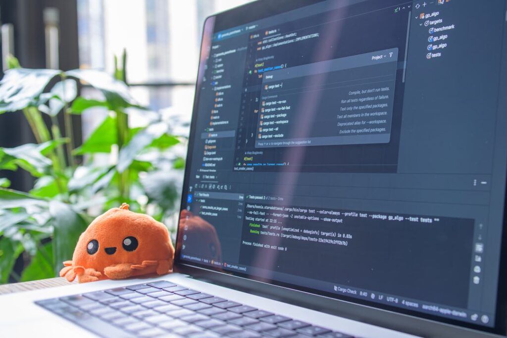 Laptop screen displaying code with a small orange plush toy representing AI assistant technology. Photo by Joel Lee