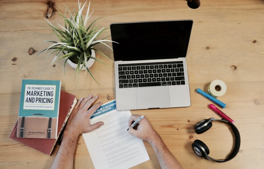 Overhead view of a marketing workspace with a laptop, notebook, marketing and pricing book, headphones, and pens — representing the cost planning involved in evaluating AI content tools