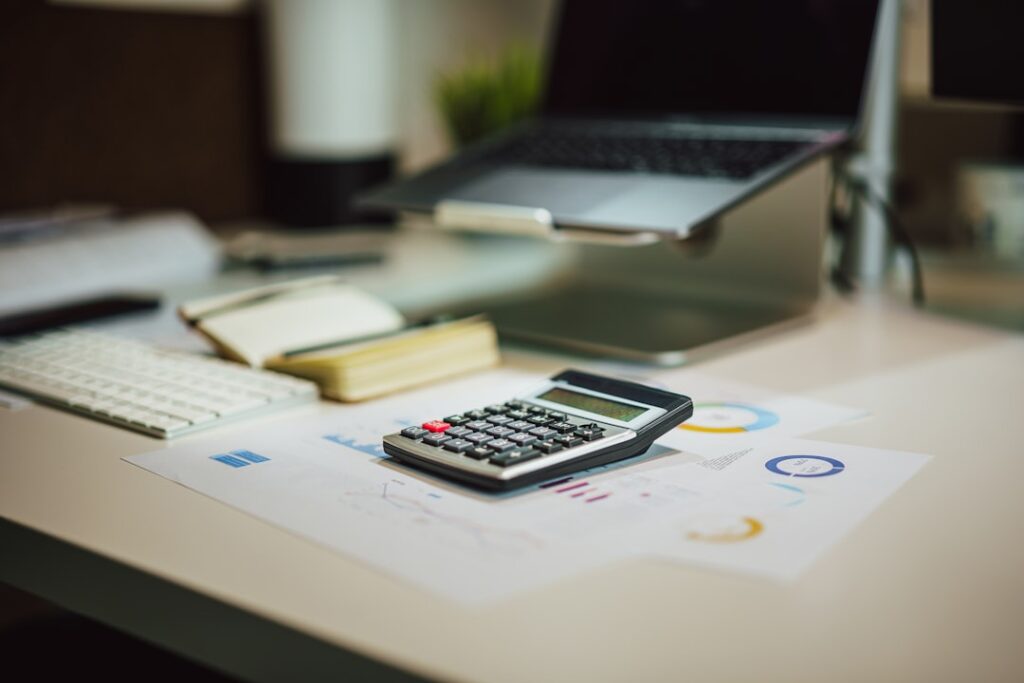 Calculator sitting on financial charts and graphs on a desk next to a laptop, representing ROI and cost analysis