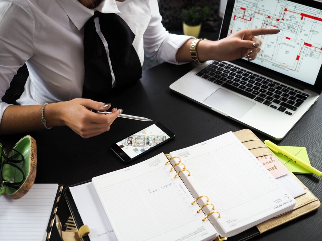 Real estate professional reviewing property floor plans on laptop with multiple devices on desk